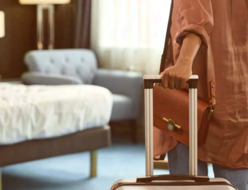 Pessoa segurando a alça de uma mala de viagem e uma bolsa marrom, em um quarto com cama e poltrona ao fundo.