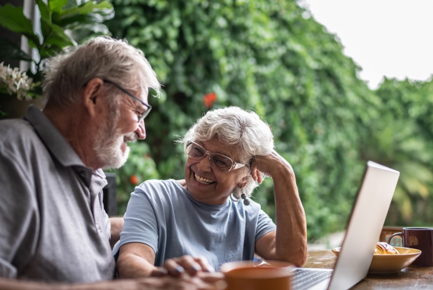 Un hombre y una mujer mayores se miran y sonríen sentados en una mesa al aire libre. Frente a ellos, una computadora portátil abierta.