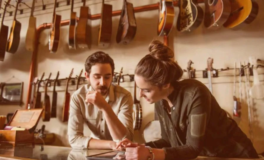 Homem e mulher em uma loja de instrumentos musicais, encostados em um balcão de vidro, olhando e interagindo com um tablet. Ao fundo, várias guitarras e instrumentos de corda estão pendurados na parede.