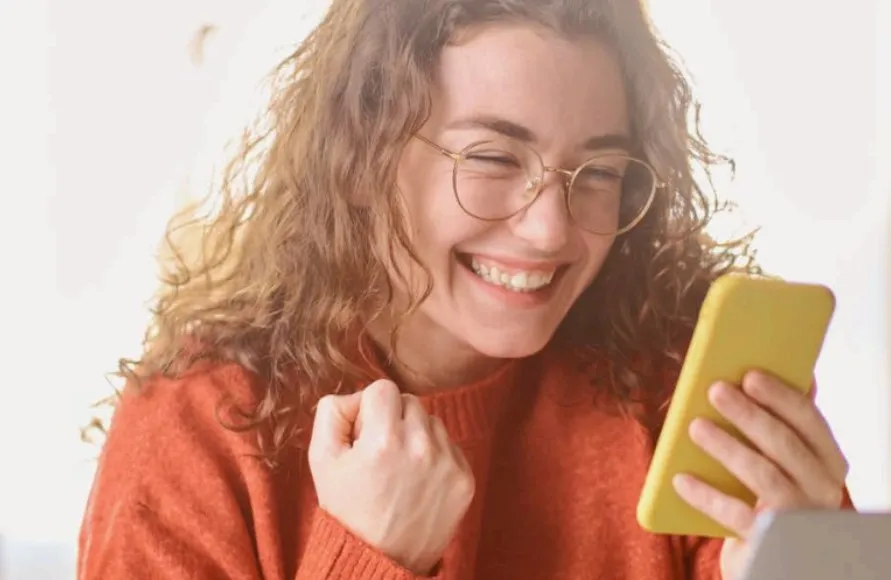 Mulher sorridente segurando um smartphone amarelo com uma mão, enquanto faz um gesto de comemoração com a outra mão, vestindo um suéter vermelho, em ambiente interno com iluminação suave ao fundo.