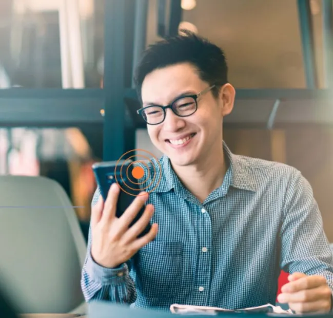 Homem jovem de óculos sorrindo enquanto segura um celular, sentado em frente a um notebook em um ambiente moderno.