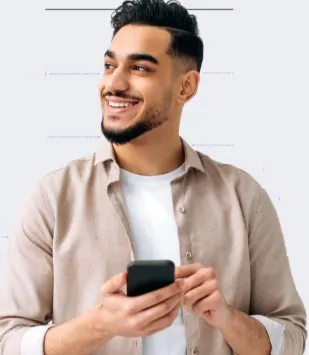 Homem jovem de barba, usando camisa bege sobre camiseta branca, sorrindo e segurando um celular enquanto olha para o lado.