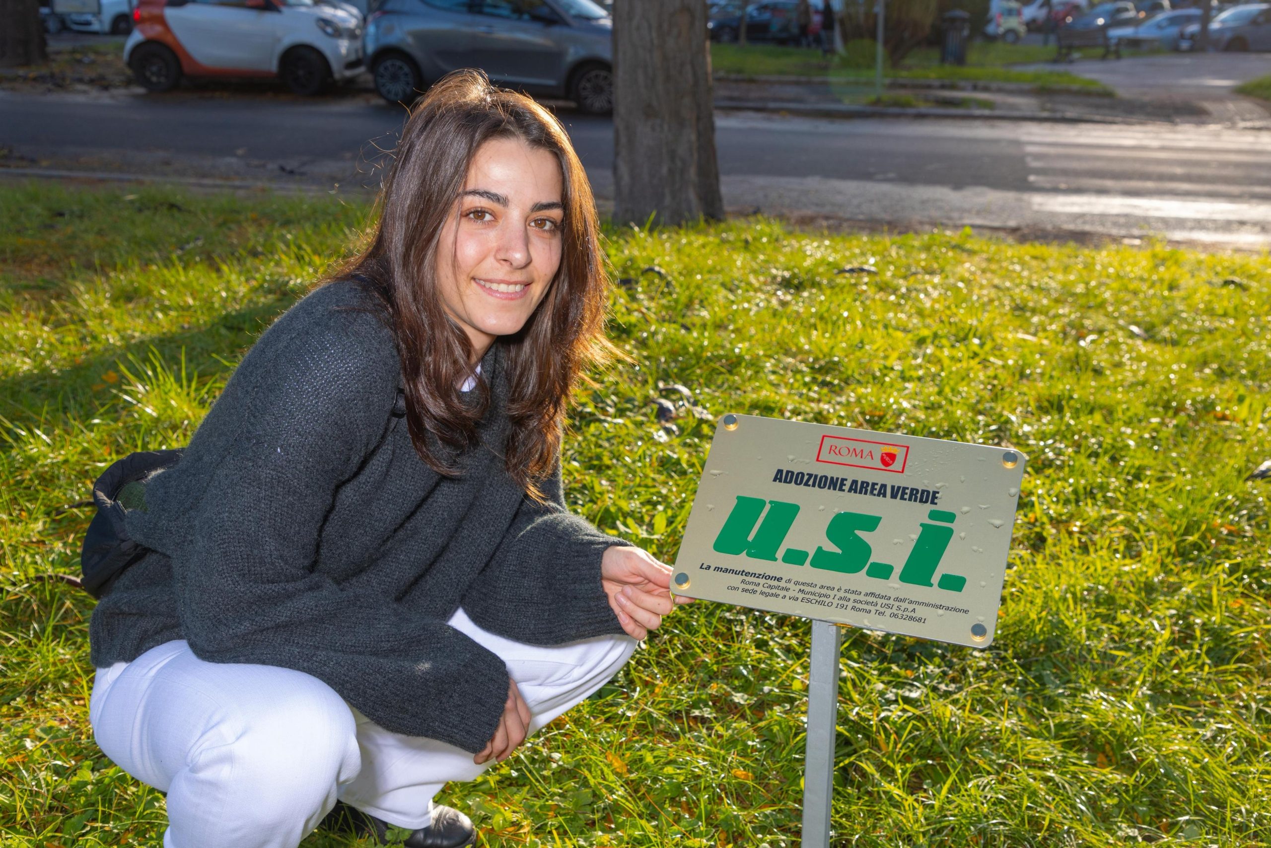 A young woman with brown hair wearing a dark gray sweater and white pants crouches on a grassy area, smiling at the camera while holding a small sign that reads “Adozione Area Verde U.S.I.” in green letters. Cars are visible in the background.