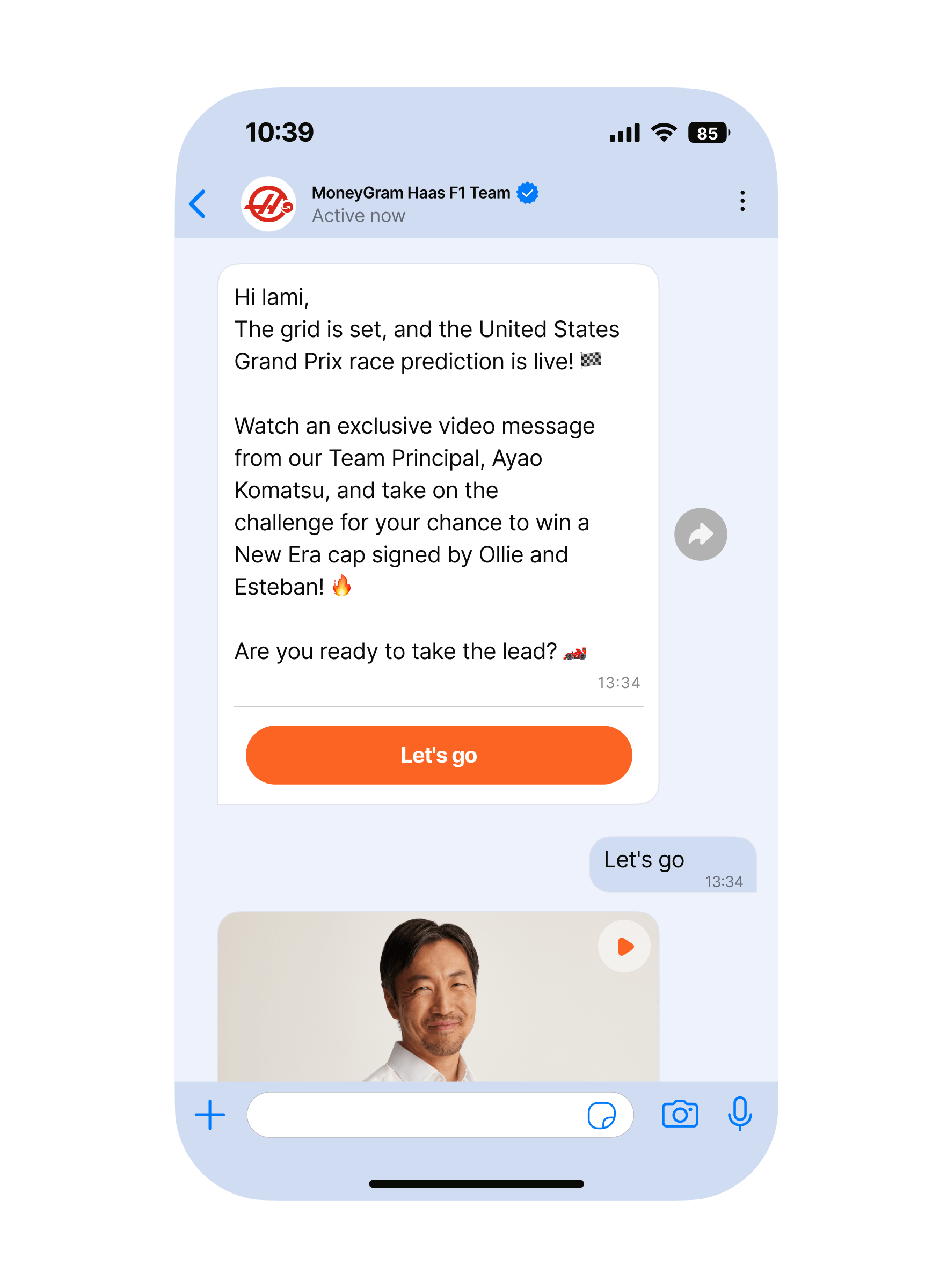 Mobile phone screen showing a light-themed messaging app conversation with the ‘MoneyGram Haas F1 Team’ at the top. A large message card invites the user to watch an exclusive video message from the team principal and join a challenge to win a New Era cap signed by Ollie and Esteban, followed by an orange ‘Let’s go’ call-to-action button. Below, a thumbnail of a video message appears above the user’s short text reply, ‘Let’s go.’