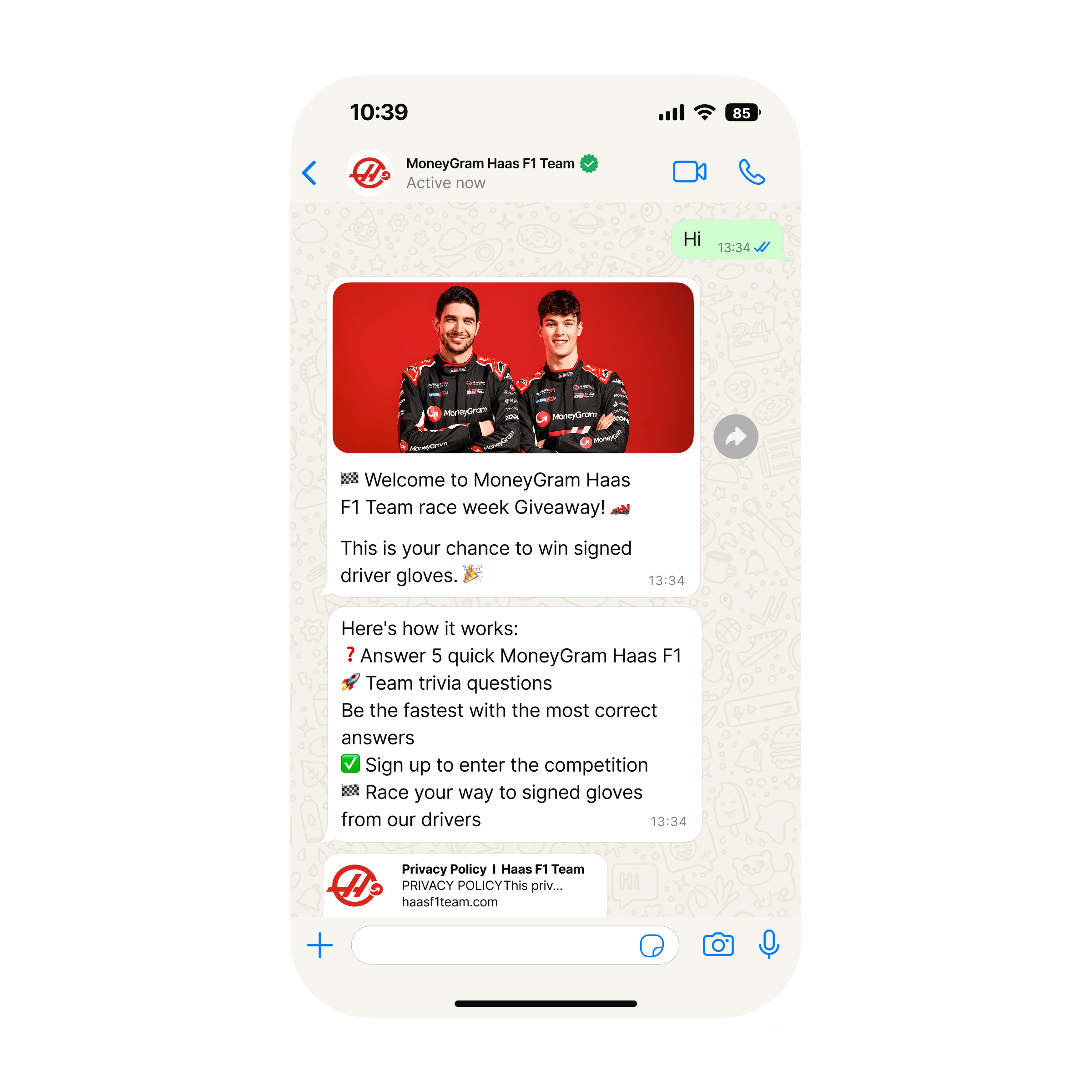 Mobile phone screen showing a WhatsApp chat with “MoneyGram Haas F1 Team.” The team has sent an image of two racing drivers in black MoneyGram Haas F1 Team suits standing in front of a bright red background. Below, messages announce a “race week Giveaway” and a chance to win signed driver gloves, followed by instructions: answer five quick MoneyGram Haas F1 Team trivia questions, be the fastest with the most correct answers, sign up to enter the competition, and race your way to signed gloves. A preview link to the team’s privacy policy website appears at the bottom of the chat.