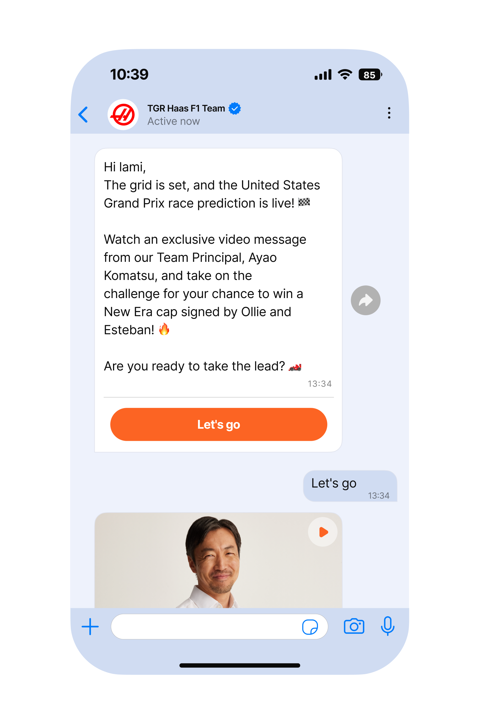 Mobile phone screen showing a light-themed messaging app conversation with the ‘TGR Haas F1 Team’ at the top. A large message card invites the user to watch an exclusive video message from the team principal and join a challenge to win a New Era cap signed by Ollie and Esteban, followed by an orange ‘Let’s go’ call-to-action button. Below, a thumbnail of a video message appears above the user’s short text reply, ‘Let’s go.’