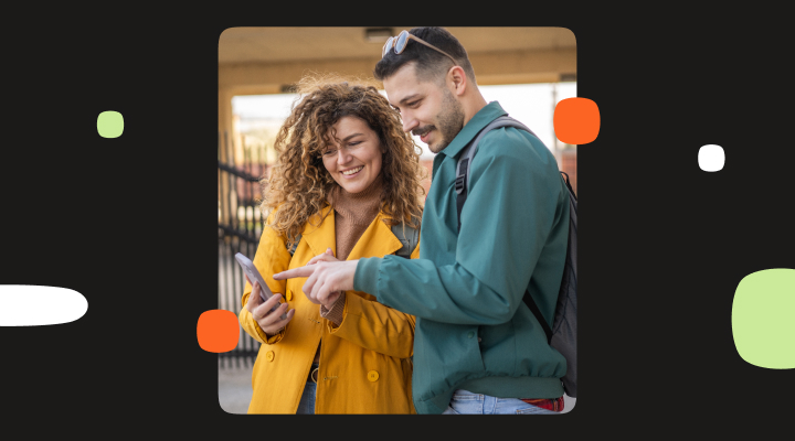 Photo of two people standing outdoors and looking at a smartphone together. One person holds the phone while the other points at the screen.