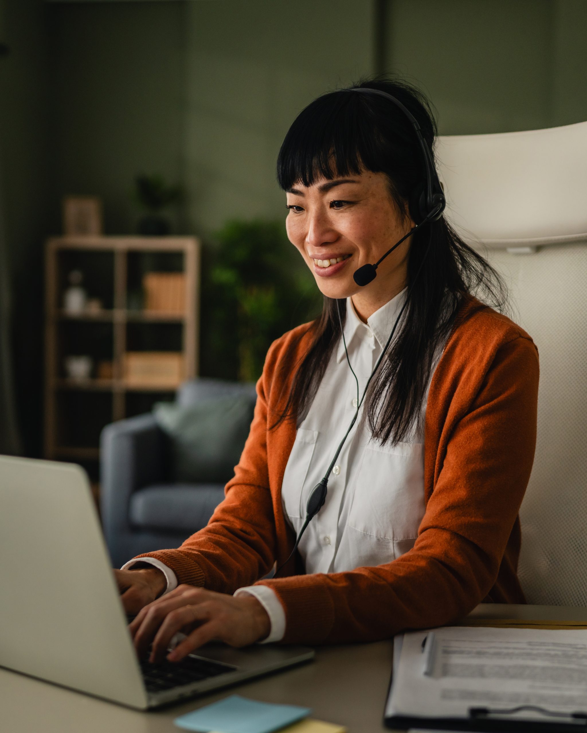 A woman wearing a headset sits at a desk, smiling while typing on a laptop. Papers, sticky notes, and a pen are on the desk, with a bookshelf and couch visible in the background.