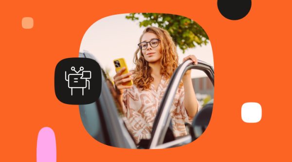 Young woman with curly hair and glasses standing beside an open car door, holding a yellow smartphone and looking at the screen. The image is framed inside a rounded shape on a bright orange background with abstract black, white, and pink design elements. A black icon showing a chatbot or automation symbol appears beside her, suggesting customer service automation or digital assistance.