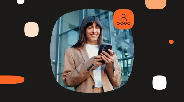 A smiling woman stands outdoors in front of a modern glass office building while looking at a smartphone she is holding with both hands. She has long dark hair with bangs and is wearing a beige blazer over a white top. The photo is framed inside a large rounded oval shape on a black background with abstract white, beige, and orange graphic elements. In the top right corner of the photo is an orange square icon showing a person above a row of connected circles, suggesting customers, audience, or user engagement.