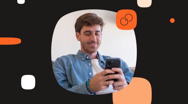 A smiling man sits indoors and looks down at a smartphone he is holding with both hands. He is wearing a light blue denim shirt over a white T-shirt. The photo is placed inside a large rounded oval frame on a black background with abstract white, beige, and orange graphic elements. In the top right corner of the photo is an orange square icon containing two linked circles, suggesting connection, integration, or customer relationship.