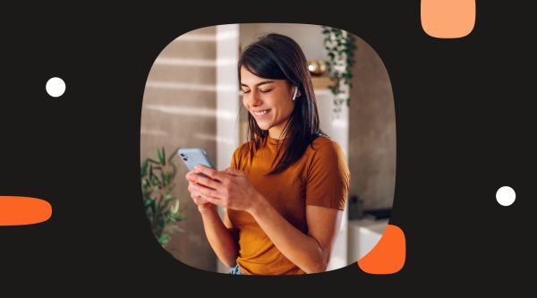 A smiling young woman with dark hair, wearing an orange t-shirt and wireless earbuds, looks down at her smartphone while typing, standing in a bright home interior with plants in the background.