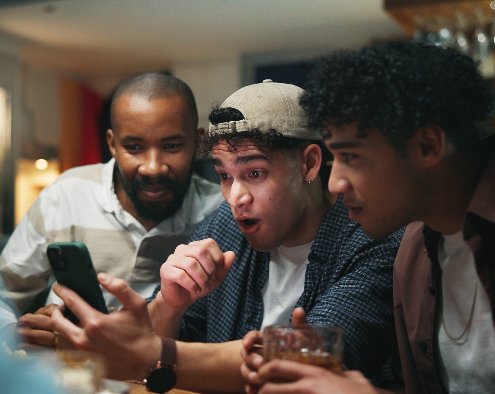 Three friends huddled together reacting with excitement to something on a smartphone during a watch party."