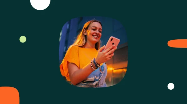 Smiling young woman in an orange t-shirt and denim overalls, wearing beaded bracelets, looks at a coral phone outdoors in warm evening light.