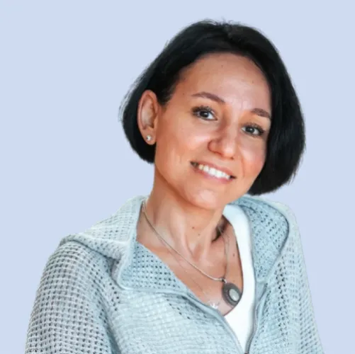 Smiling woman with short dark hair wearing a light blue textured zip-up jacket and a necklace against a light blue background
