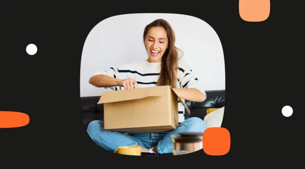 A smiling woman opens a cardboard delivery box while seated on a sofa.
