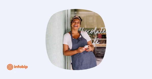 Smiling cafe employee in apron leaning by storefront while holding a smartphone