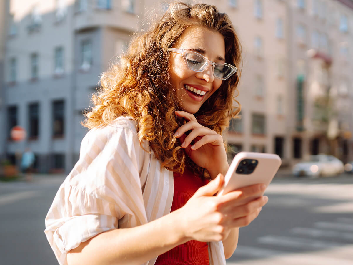 Woman standing on a city street in sunlight, looking at her smartphone.