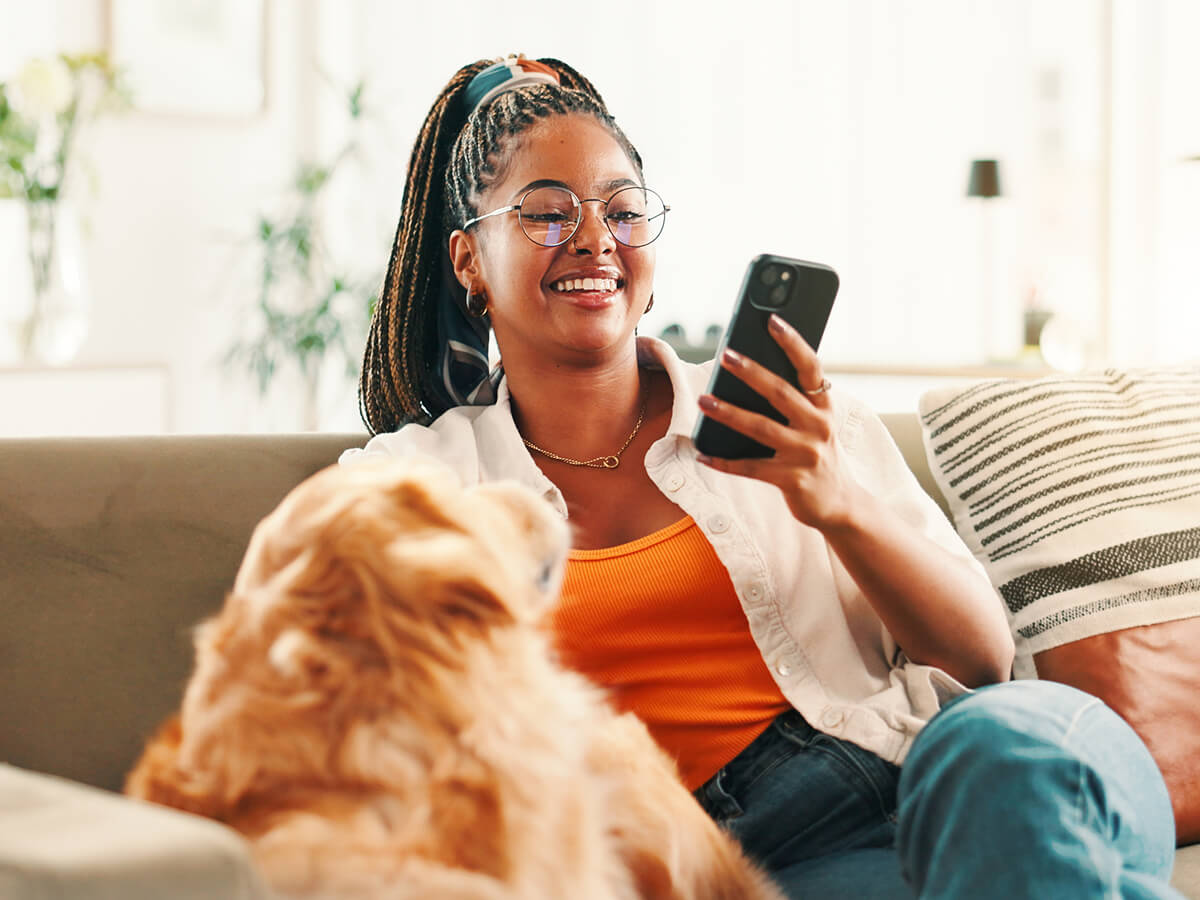 Woman relaxing on a sofa and checking her phone while a dog sits nearby.