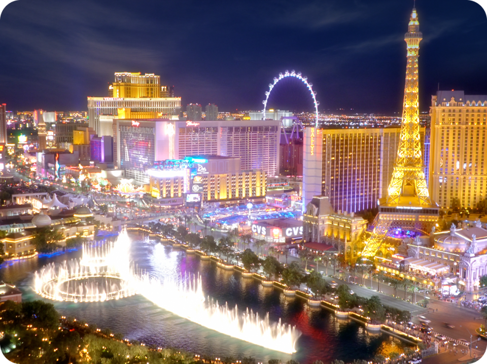 This image represents a nighttime aerial view of the Las Vegas Strip featuring illuminated landmarks including the Eiffel Tower replica at Paris Las Vegas, Bellagio Hotel and Casino, a large observation wheel, and vibrant lights from surrounding buildings. The Bellagio Fountain displays a water show in the foreground