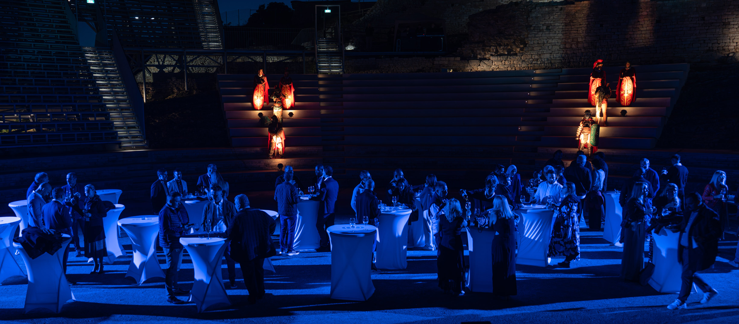 Evening networking reception in an outdoor amphitheater, guests mingling around tall cocktail tables under blue lighting, with Roman-era costumed performers illuminated on the tiered steps.