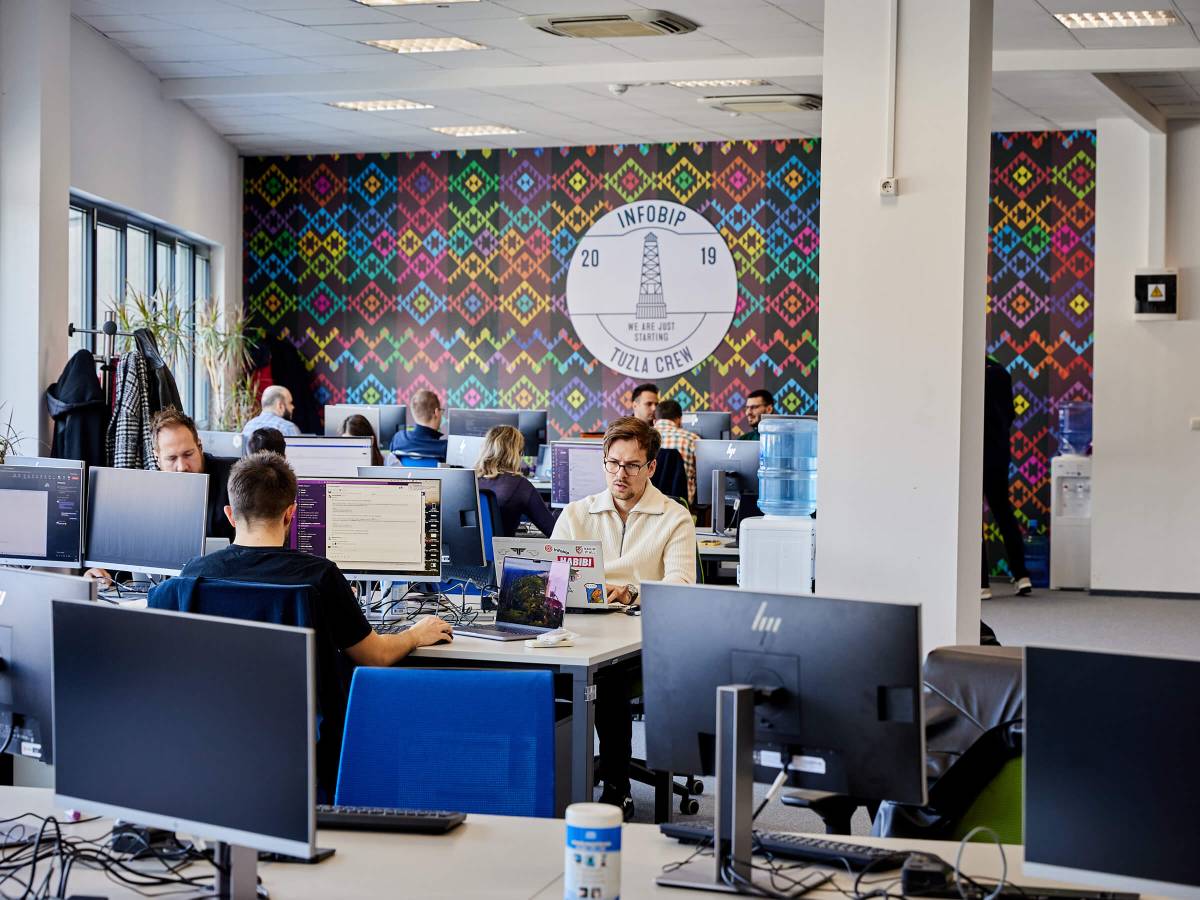 Open‑plan office with colorful Infobip Tuzla Crew wall mural and staff working at computer stations