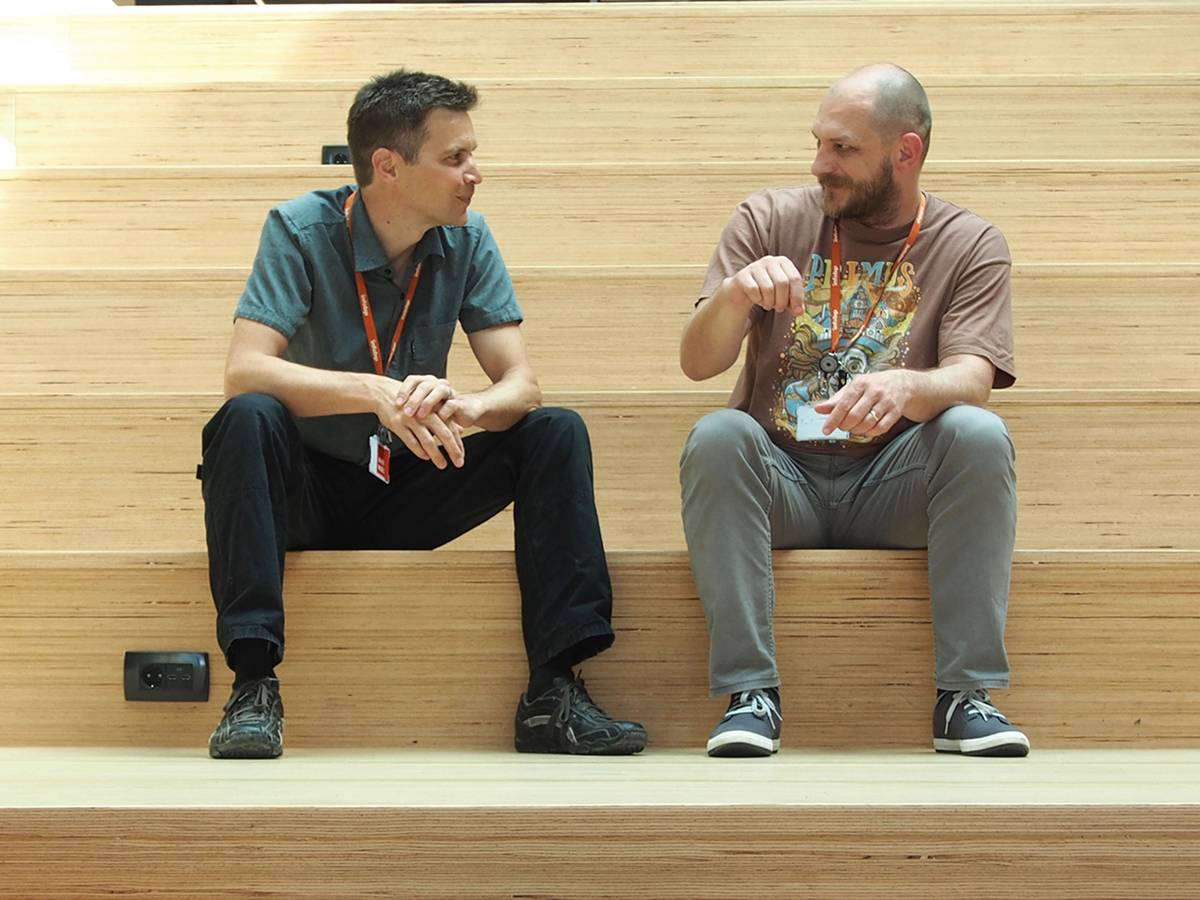 Two colleagues having a casual discussion on wooden tiered seating in the office, wearing company lanyards